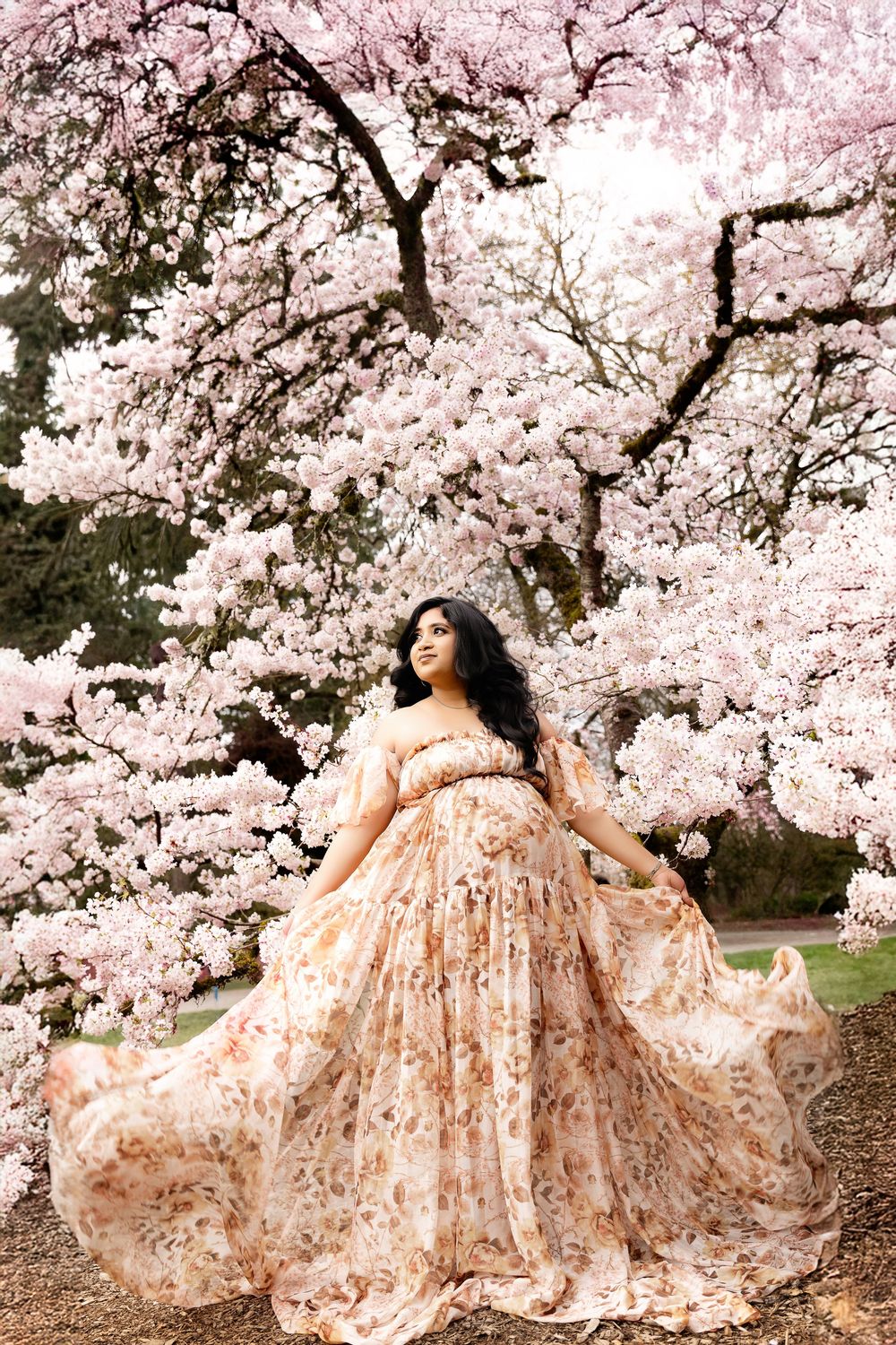Pregnant woman in floral gown poses under blooming cherry blossom tree in spring garden.