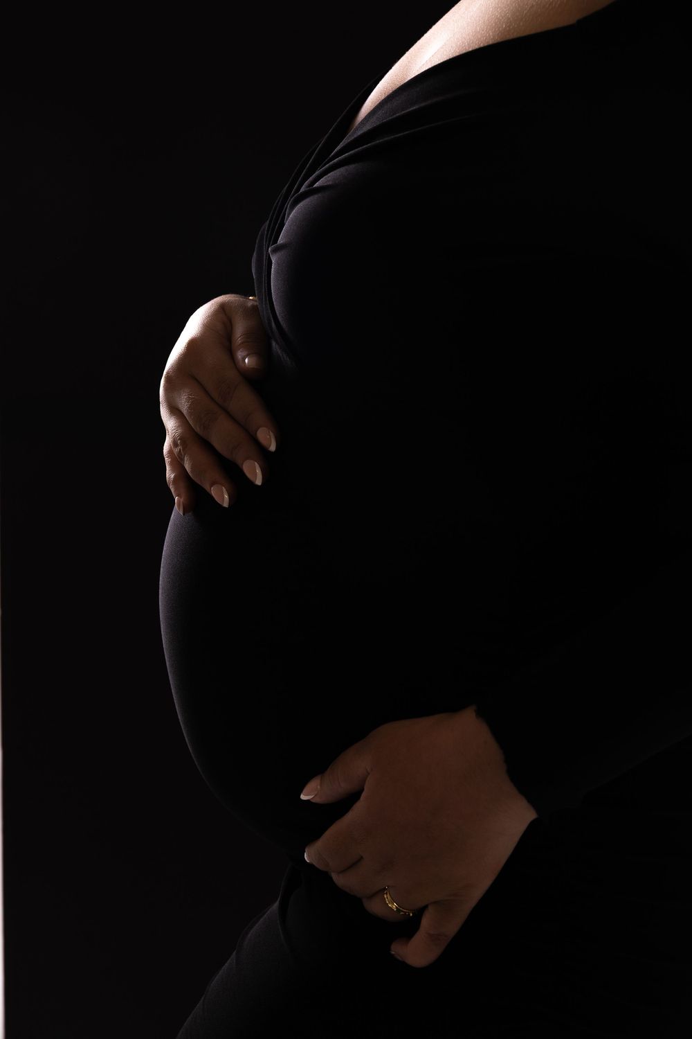 Silhouette of a pregnant woman in black clothing cradling her baby bump against a dark background.