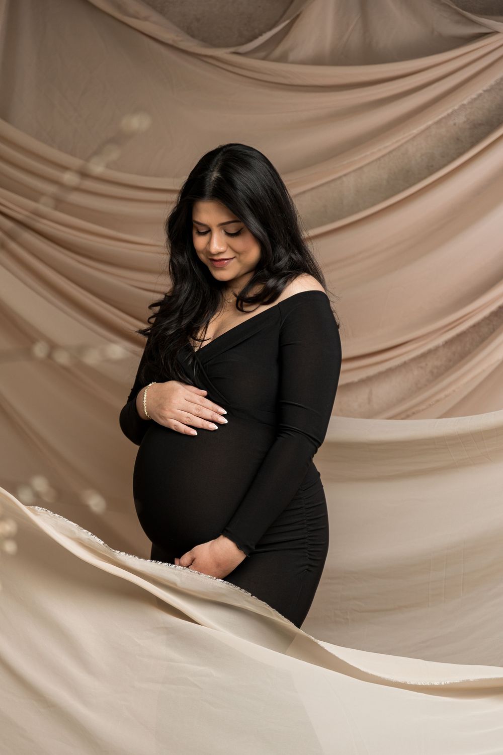 Pregnant woman in black dress cradling baby bump, smiling softly against flowing beige fabric backdrop.
