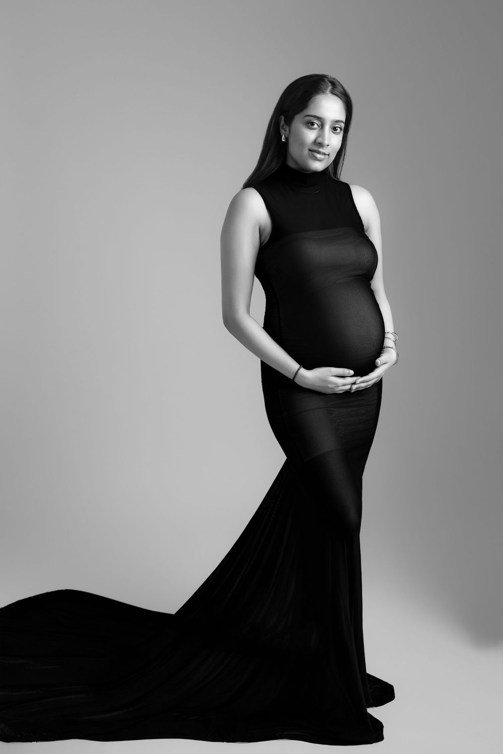 Pregnant woman in elegant black gown cradling baby bump, studio maternity portrait in black and white.