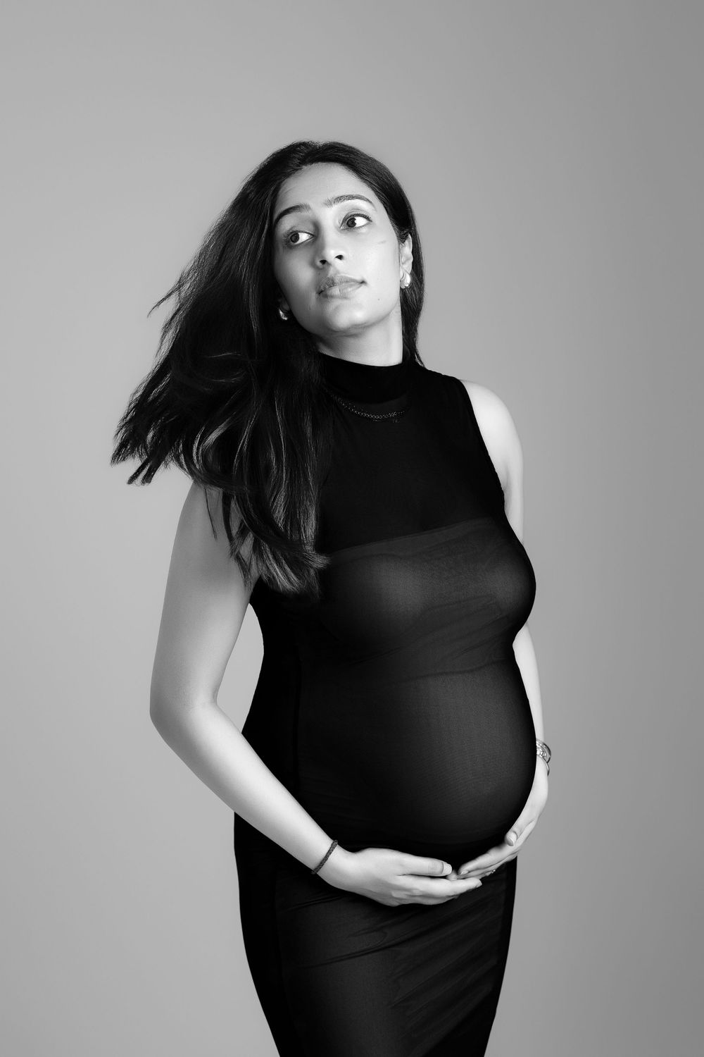 Black and white portrait of a pregnant woman in a black dress cradling her baby bump in a studio setting.