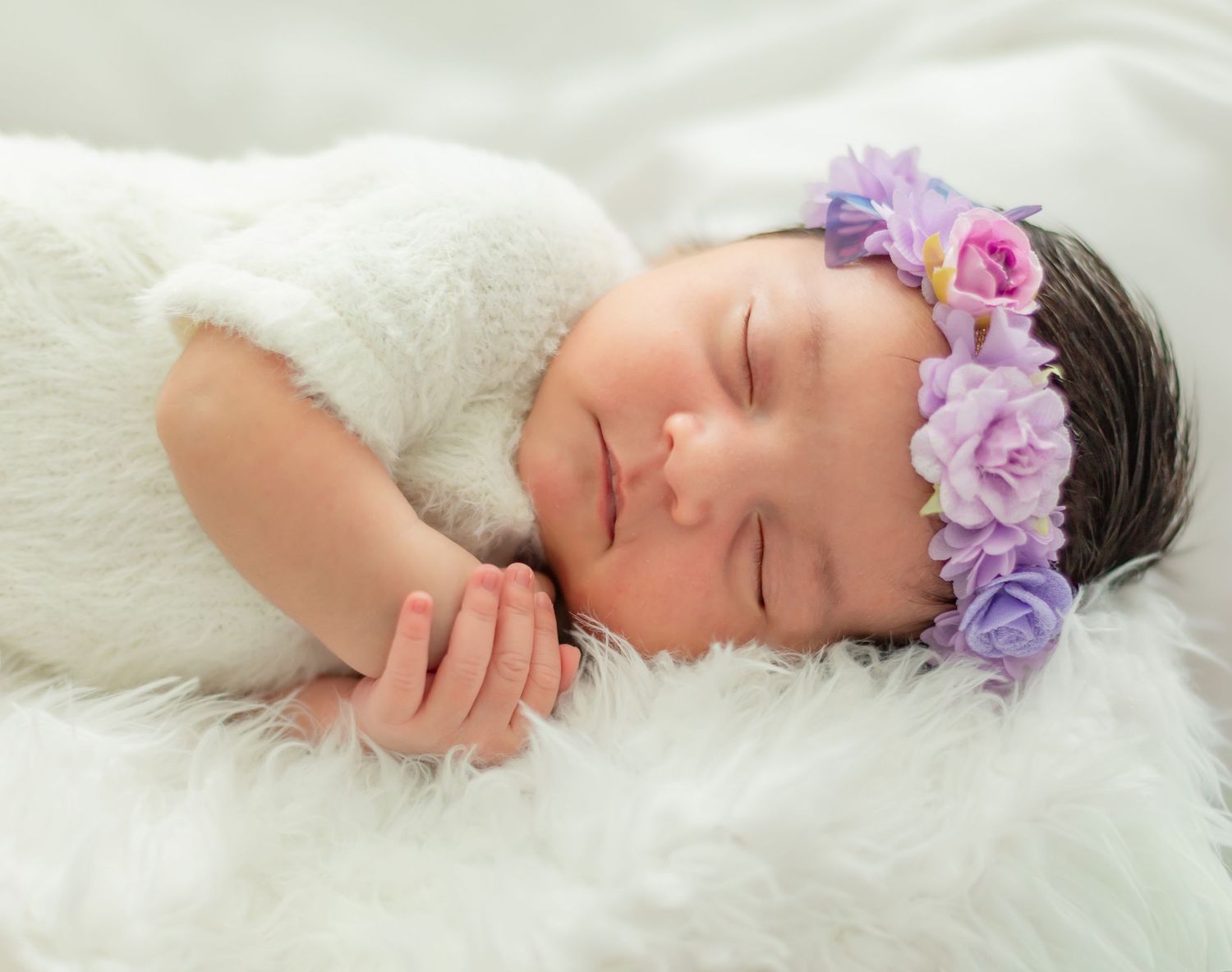 Peaceful newborn baby girl sleeping on white fur, wearing a delicate purple floral headband and white outfit.