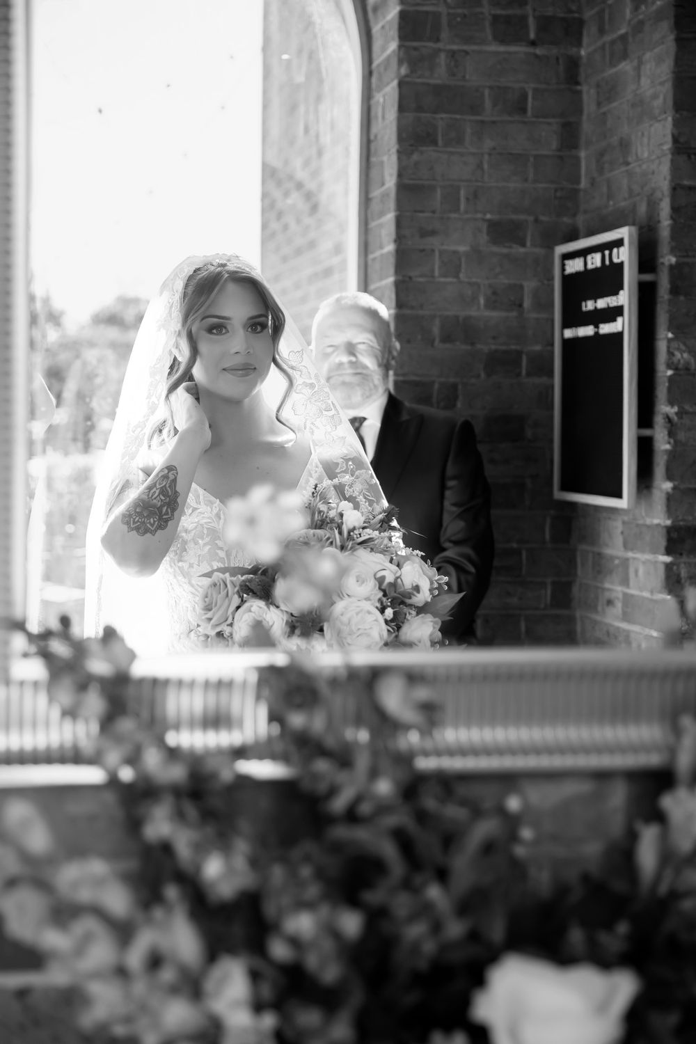 Black and white photo of a bride holding a bouquet with her father outside a brick church on her wedding day.