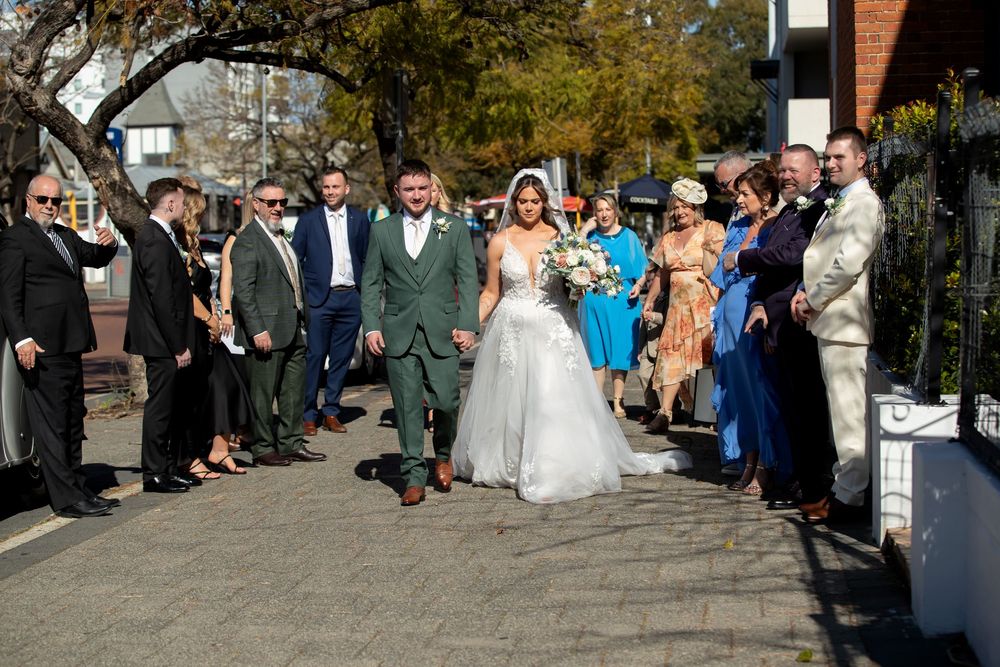 Bride and groom walk through a wedding party lineup outdoors on a sunny day, surrounded by guests in formal attire.