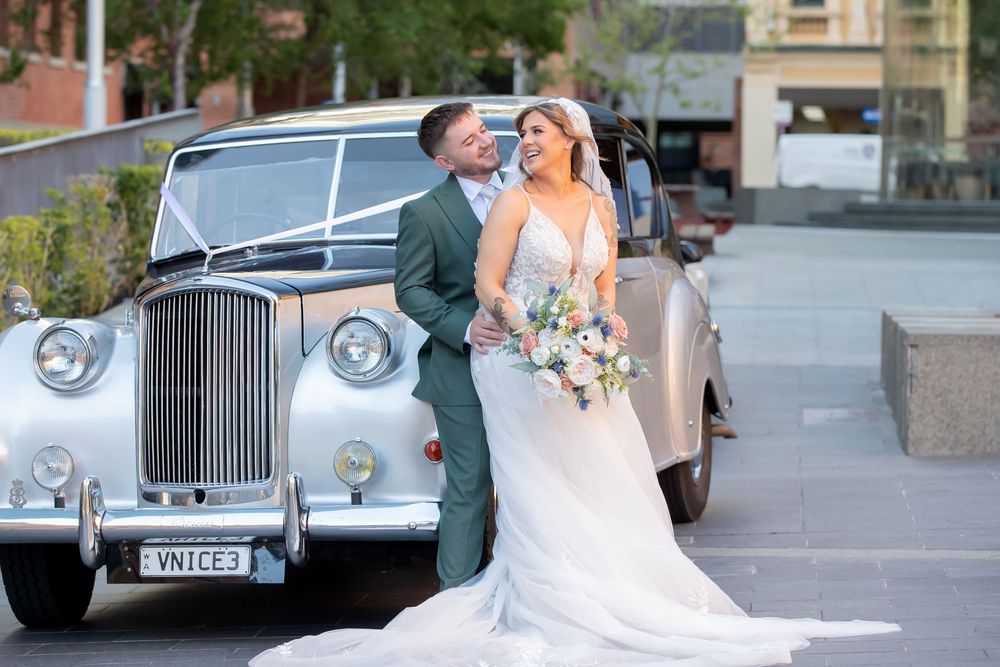 Bride and groom posing beside a vintage silver wedding car, bride holding a pastel bouquet in an elegant white gown.