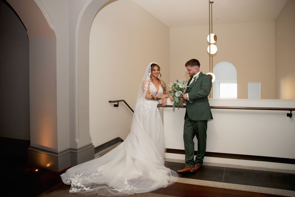 Bride in white gown and groom in green suit share a tender moment on elegant staircase during wedding ceremony.