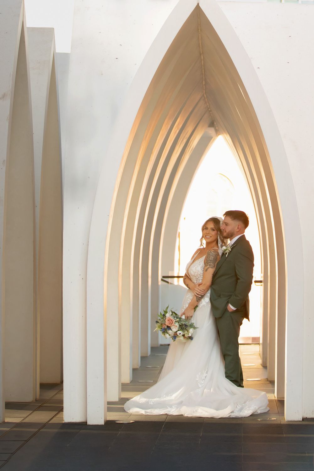 Bride and groom embracing under elegant white Gothic arches, bride holding floral bouquet in a stunning wedding portrait.