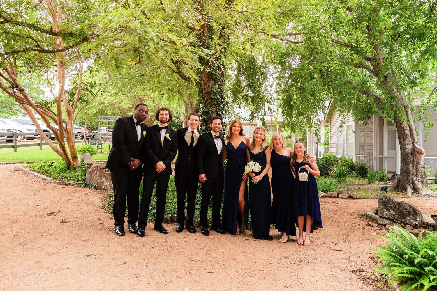 Wedding party in black attire posing outdoors under lush green trees on a sandy path near a rustic building.