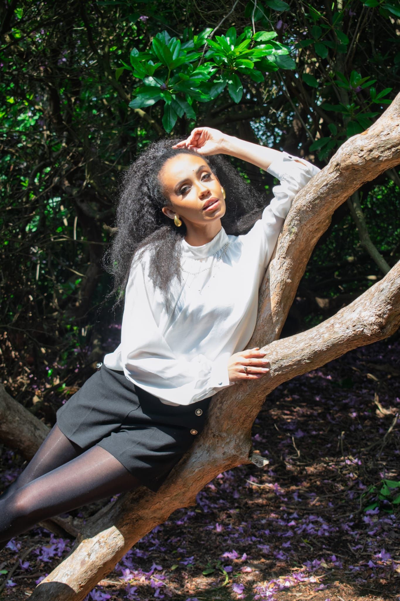 Young Black woman with curly hair in white blouse and black skirt leaning on a tree branch outdoors.