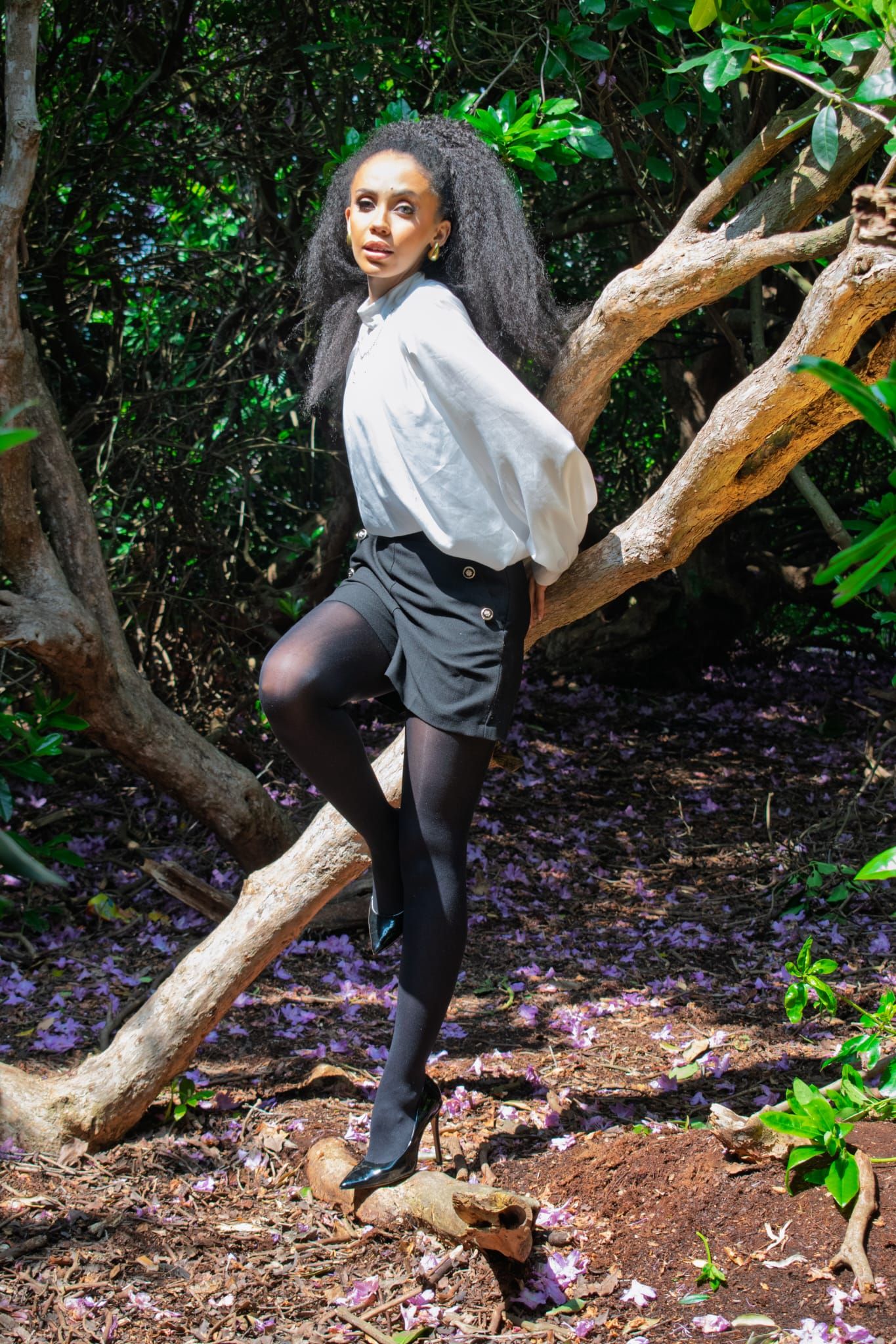 Stylish Black woman with voluminous curly hair posing in white blouse and black mini skirt in a forest.