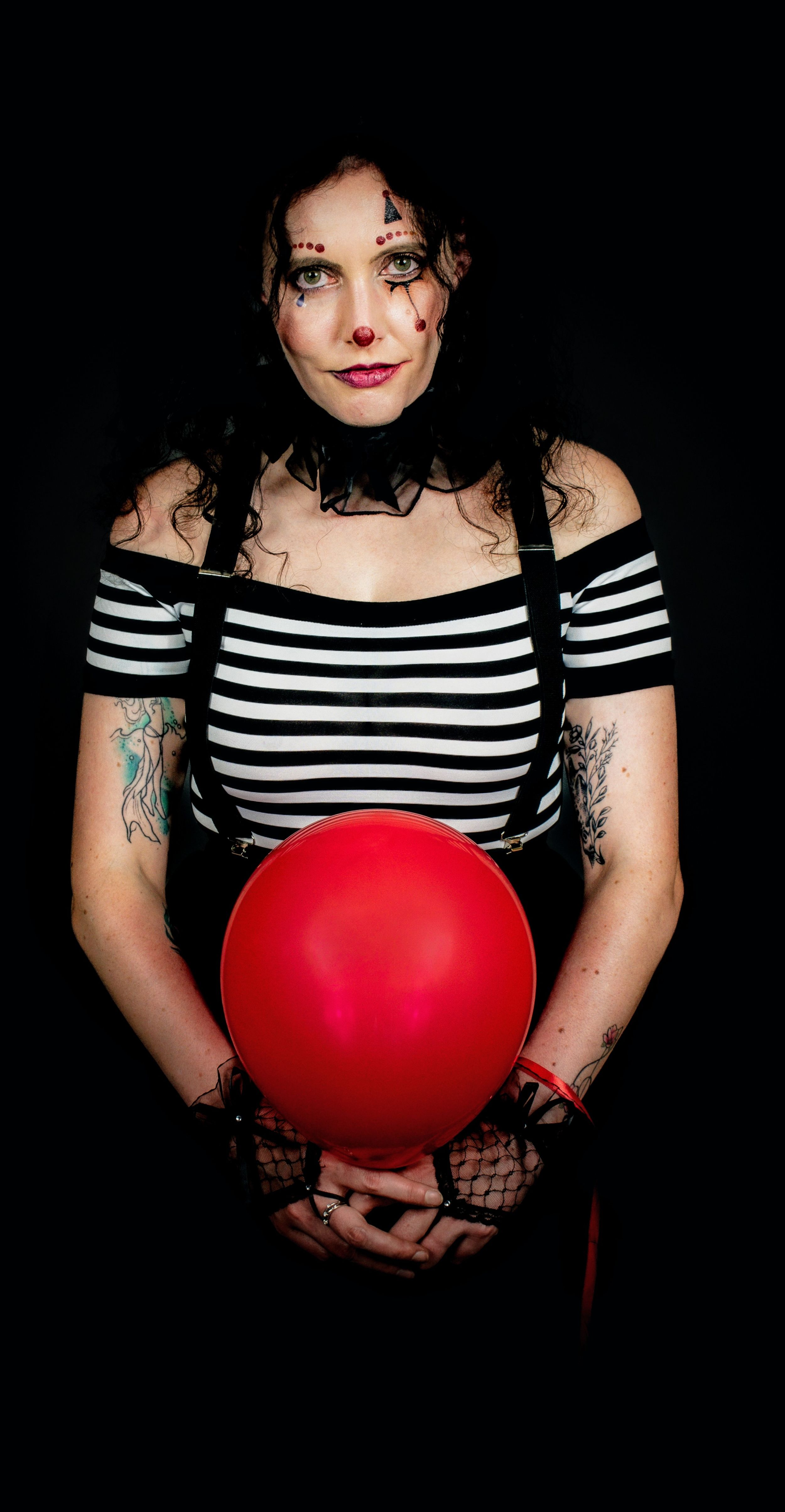 Woman in Halloween clown makeup holding a red balloon, wearing striped top with tattoos on dark background.