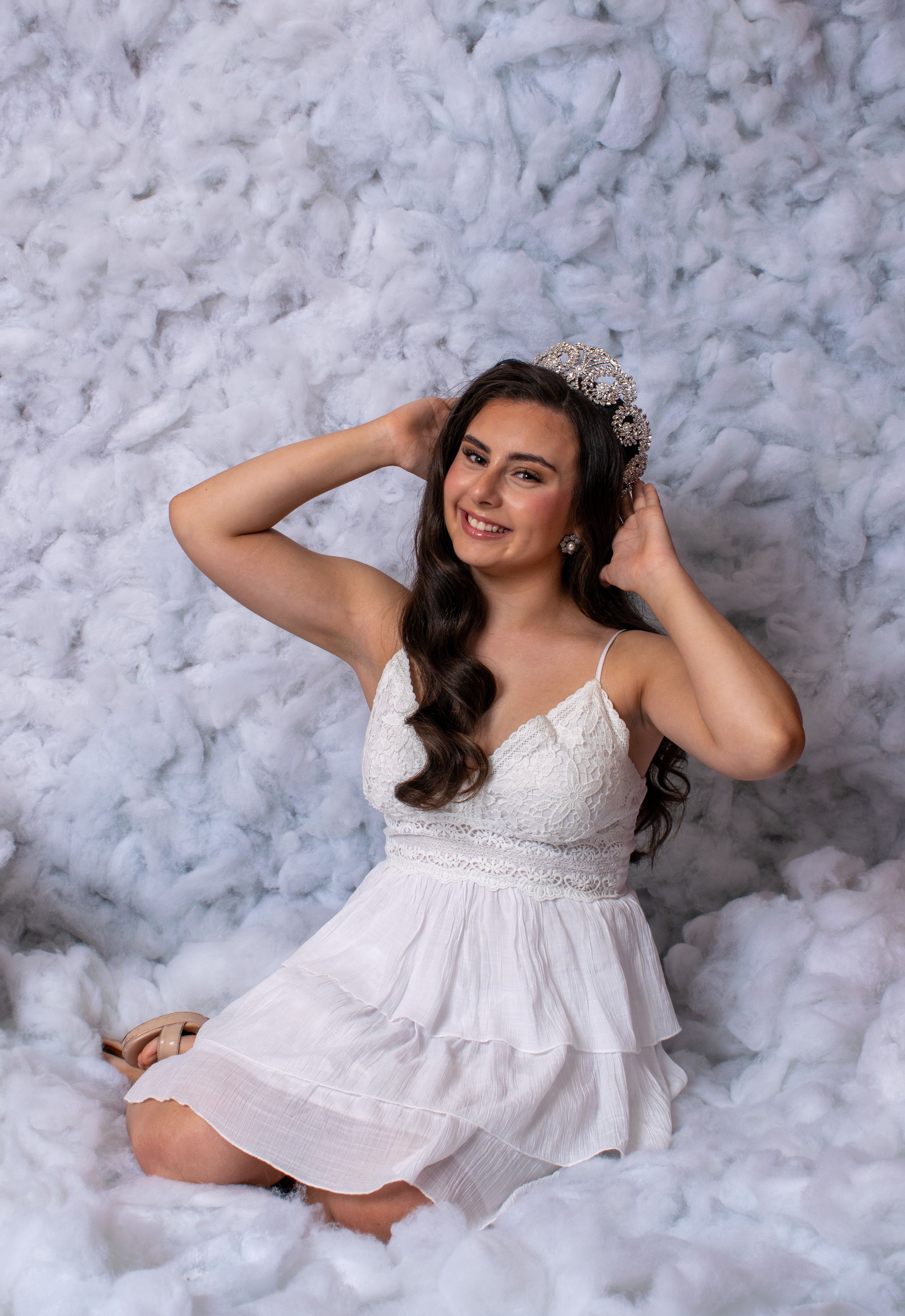 Smiling brunette teen wearing a white dress and tiara, posing against a soft white feathery backdrop.