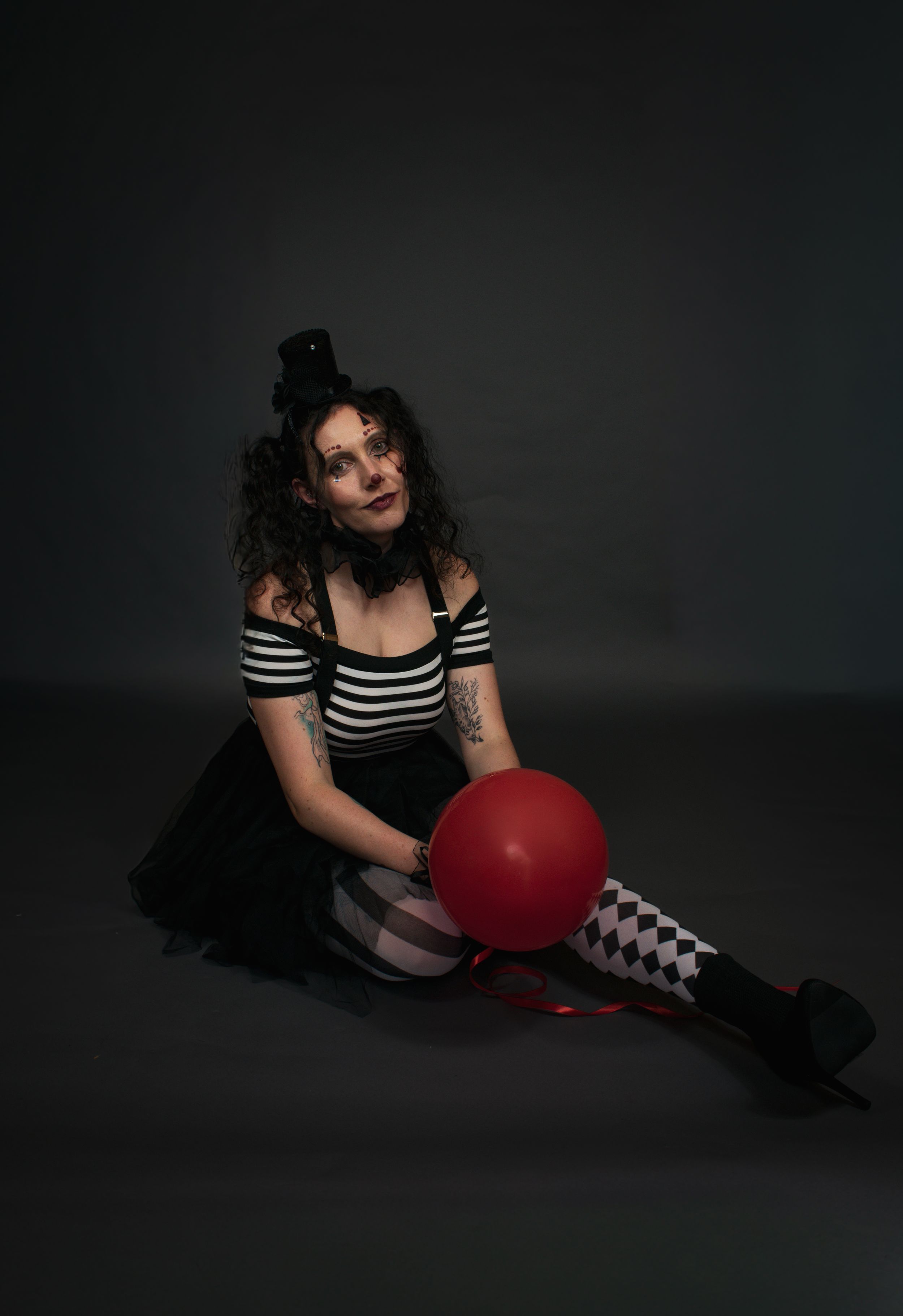 Woman in dark clown costume with red balloon sitting on floor against gray background in full body shot.
