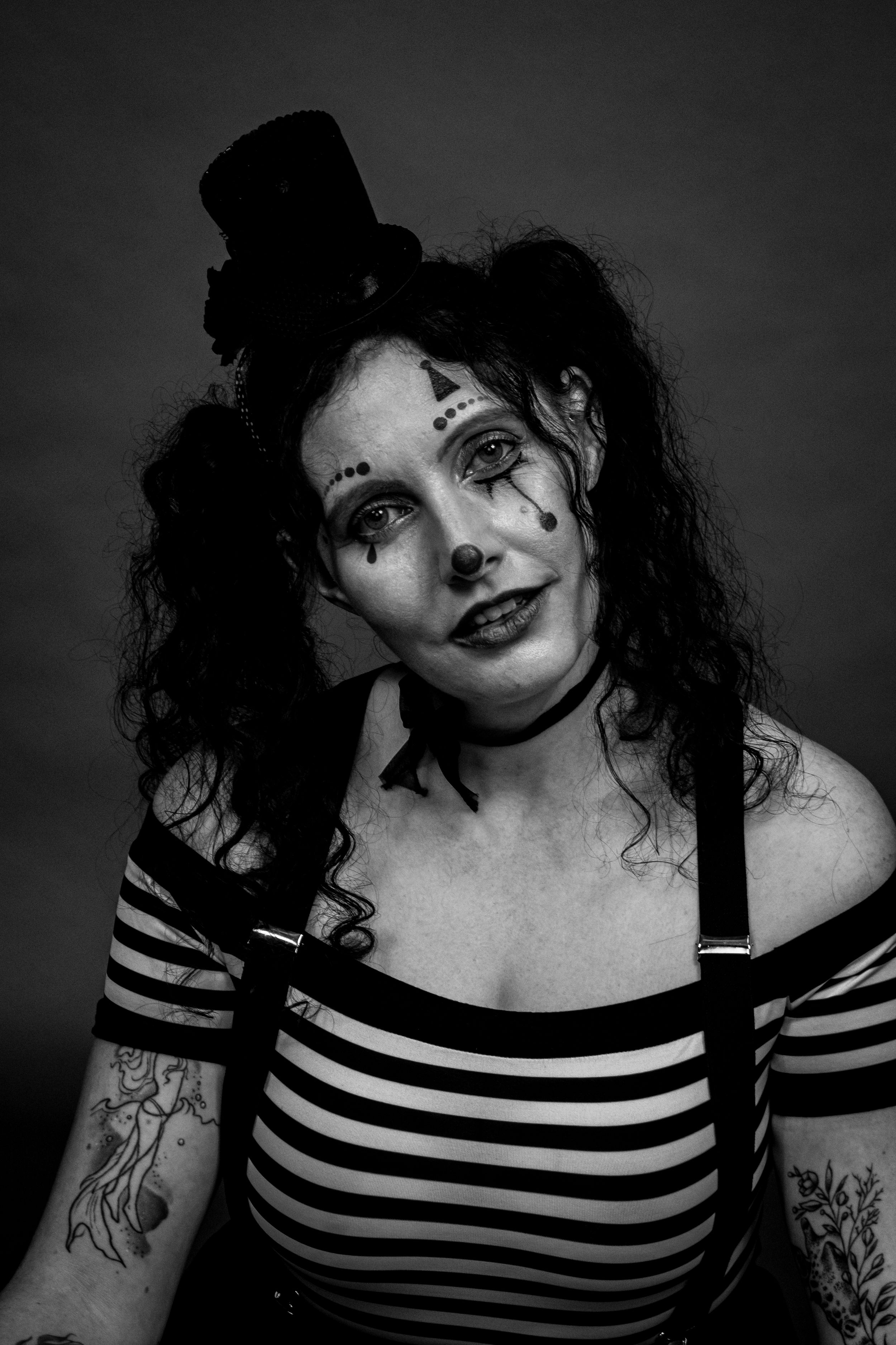 Black and white portrait of woman in theatrical clown makeup wearing striped top with suspenders and tattoos.