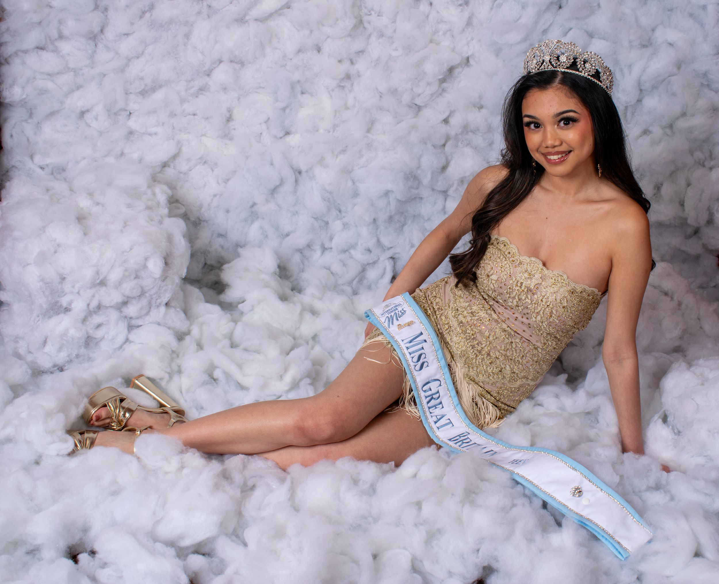 Young woman in gold pageant dress wearing a crown and sash, posing on a white fluffy background.