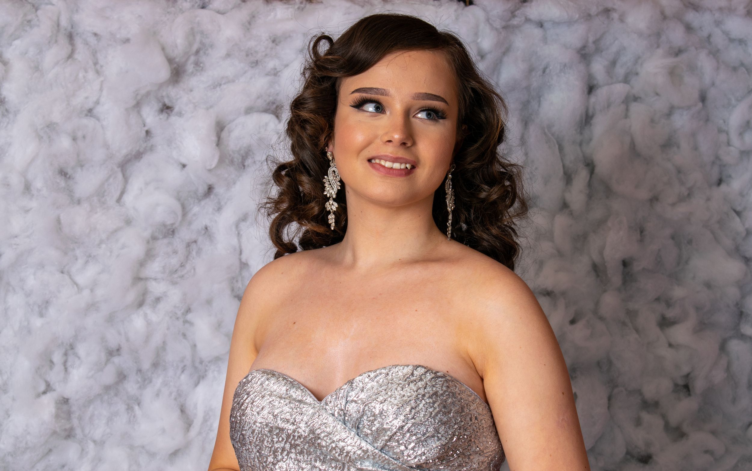 Teen girl in silver sequin strapless gown with curly hair and drop earrings posing for formal portrait.