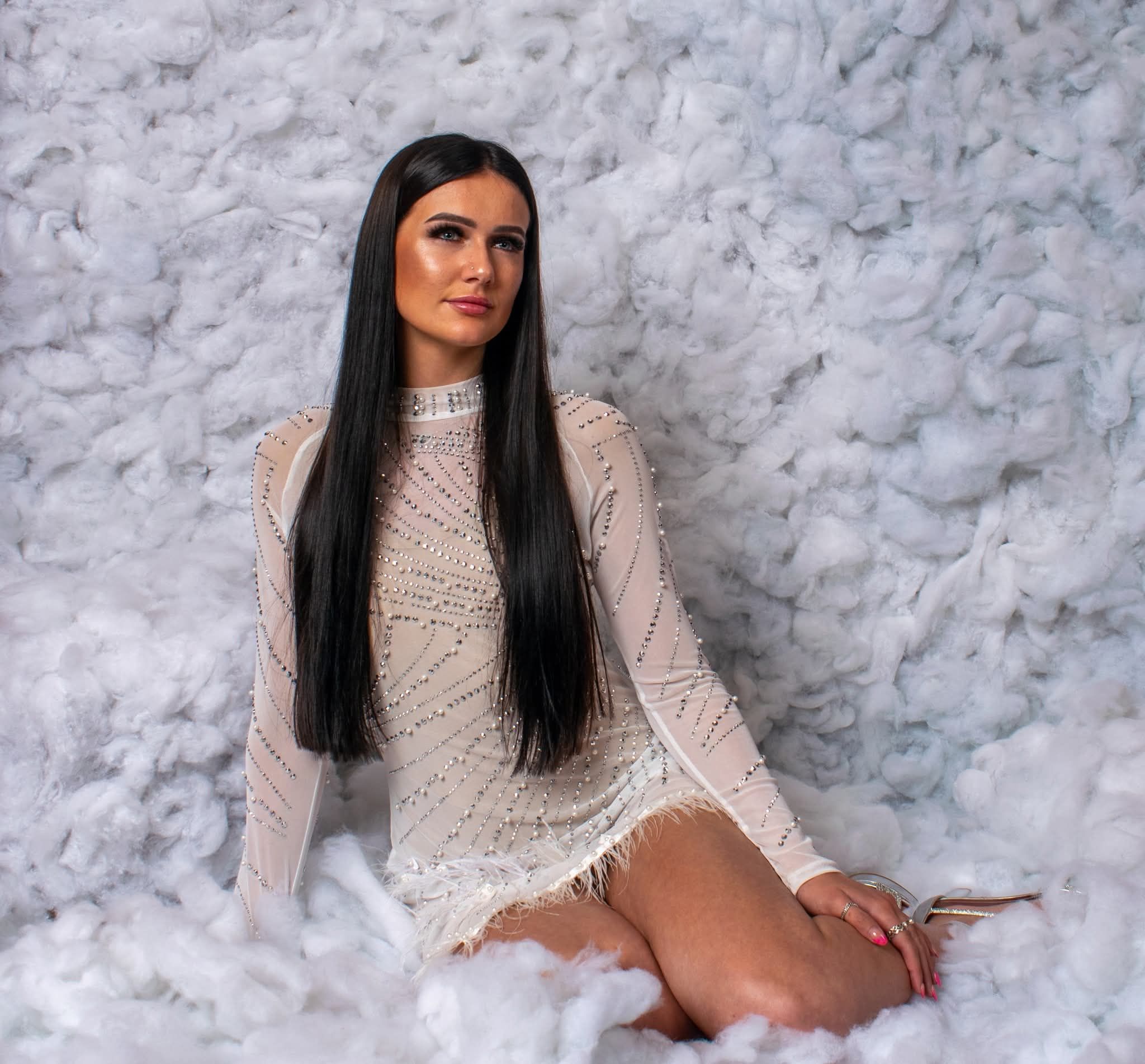 Young woman with long dark hair wearing a white lace mini dress, posing against a fluffy white background.