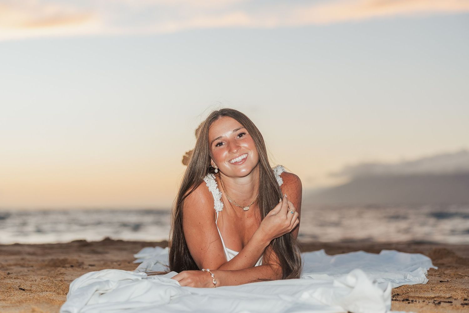 Smiling brunette woman lies on white blanket on sandy beach at sunset.