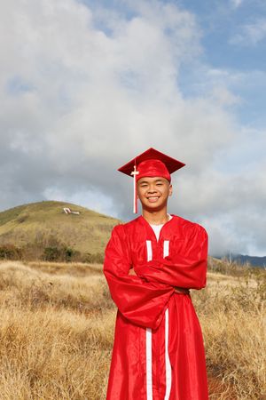 Smiling graduate in red cap and gown poses with crossed arms in scenic outdoor field.