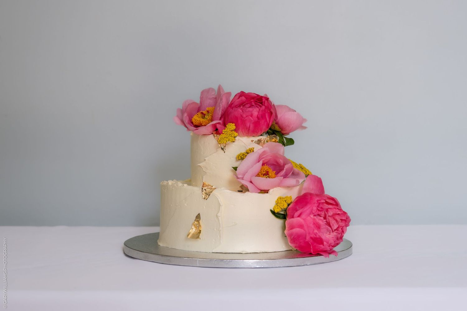 Two-tier white buttercream wedding cake decorated with vibrant pink peonies and gold leaf accents on a silver cake board.
