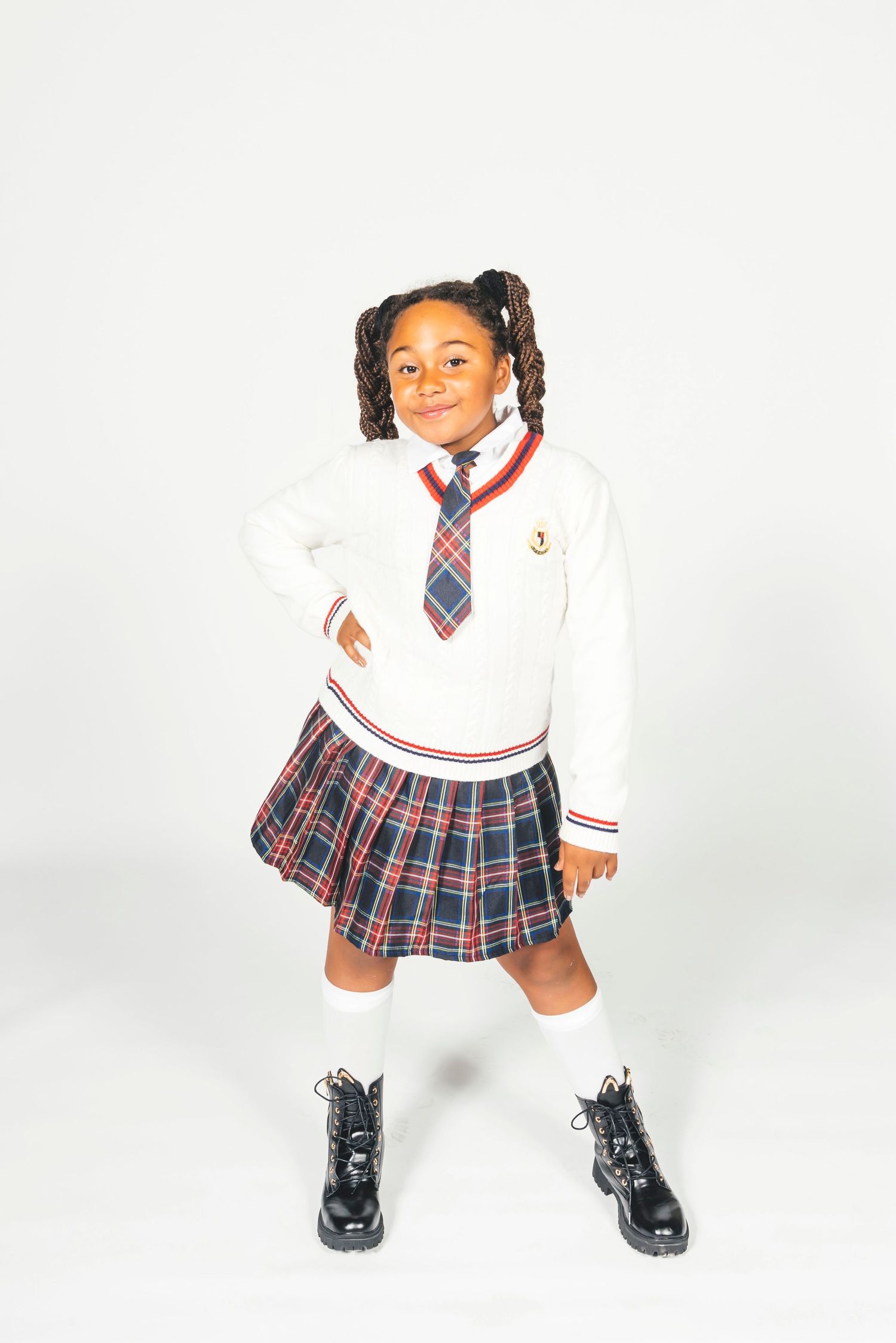 Smiling girl wearing plaid school uniform, white sweater with emblem, tie, knee-high socks, and black boots.