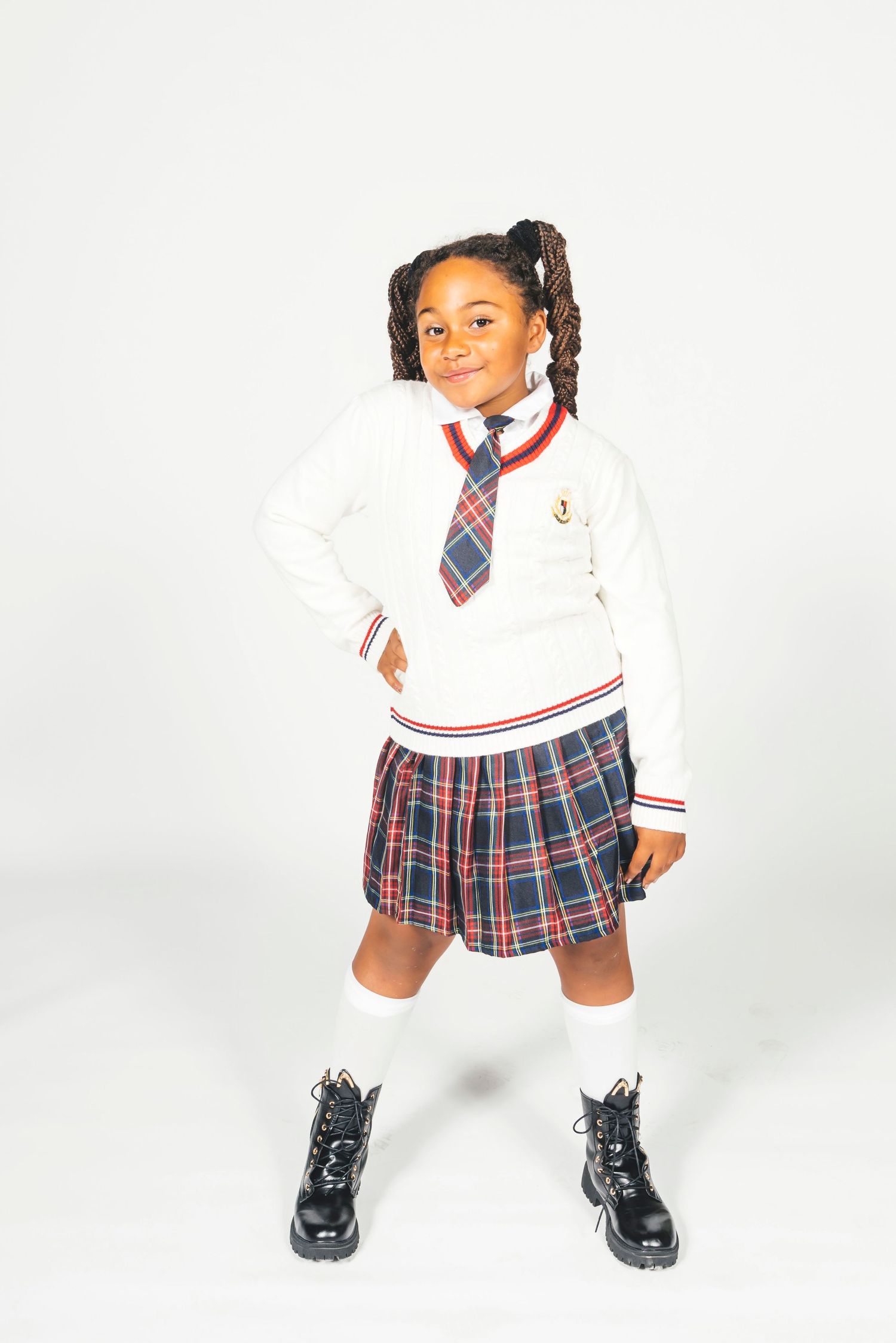 Young girl in plaid school uniform with white sweater, tie, knee socks, and black boots posing confidently.
