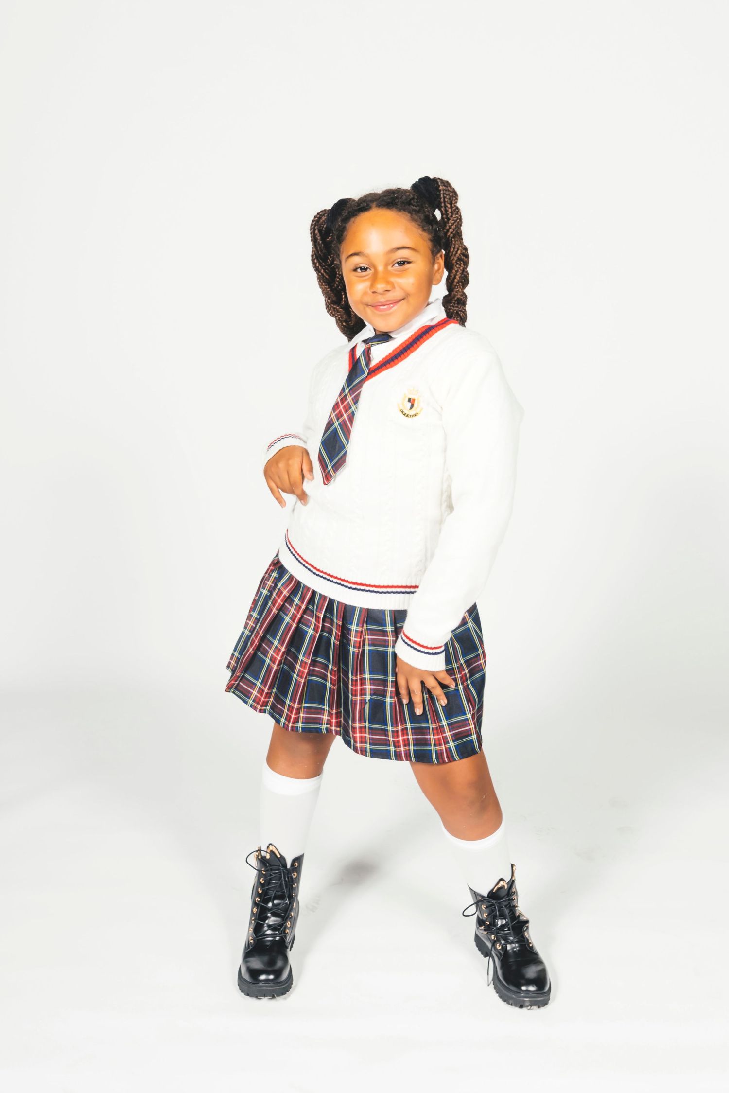 Young girl in school uniform with plaid skirt, white sweater, tie, knee socks, and black boots, smiling confidently.