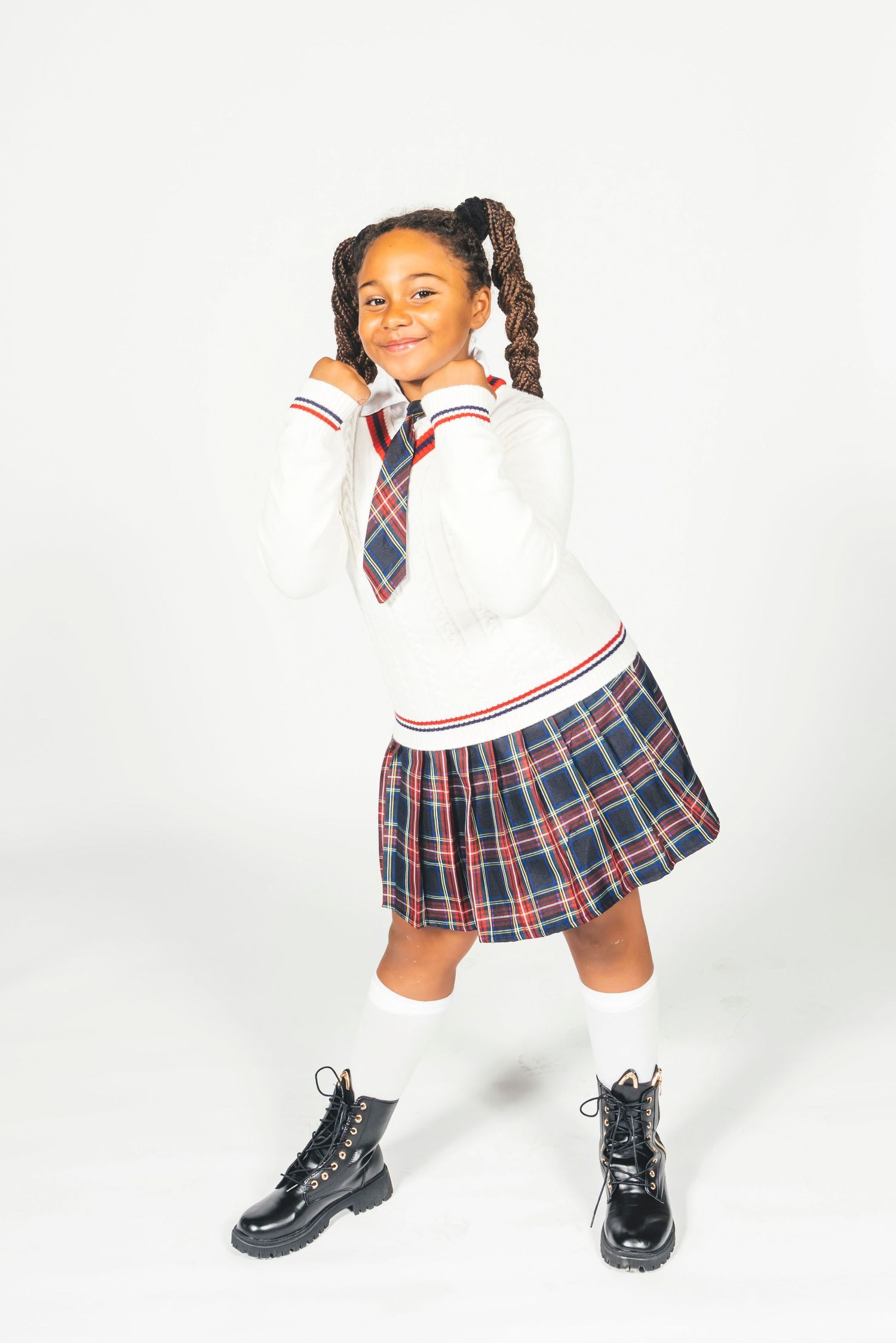 Cheerful girl in plaid school uniform and white sweater holding her tie, wearing knee socks and black boots.
