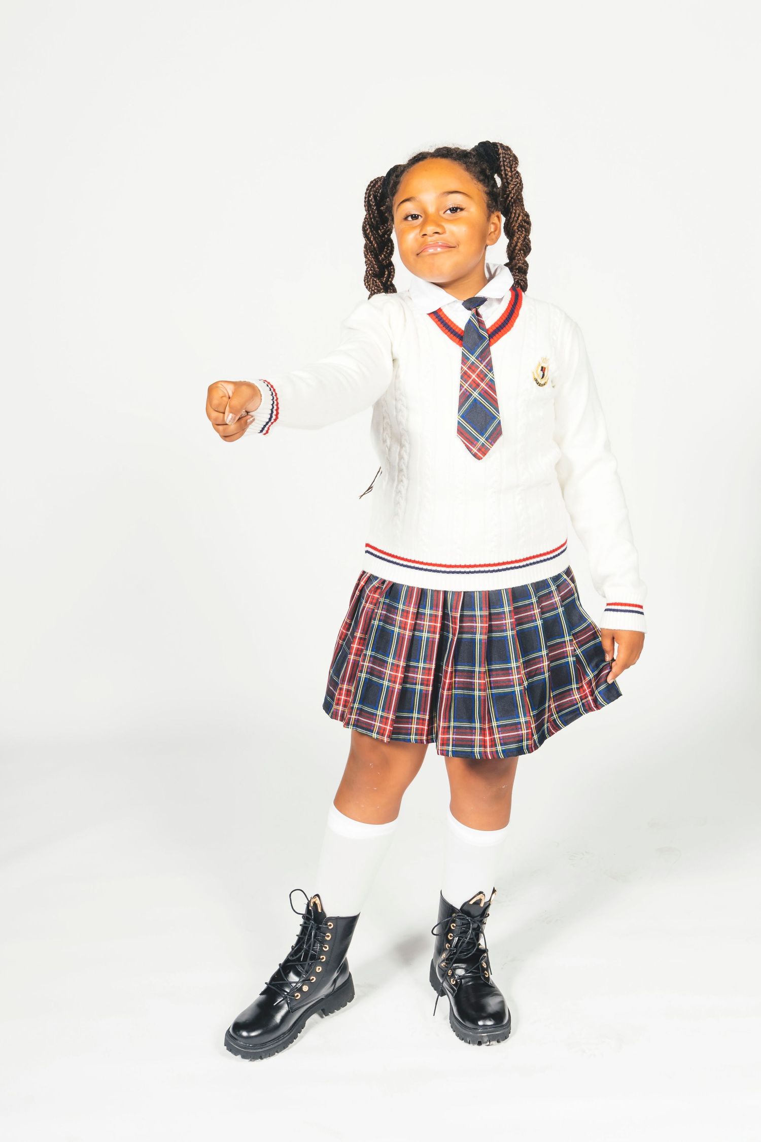 Young girl in school uniform with plaid skirt, white sweater, tie, knee socks, and black boots, pointing forward.
