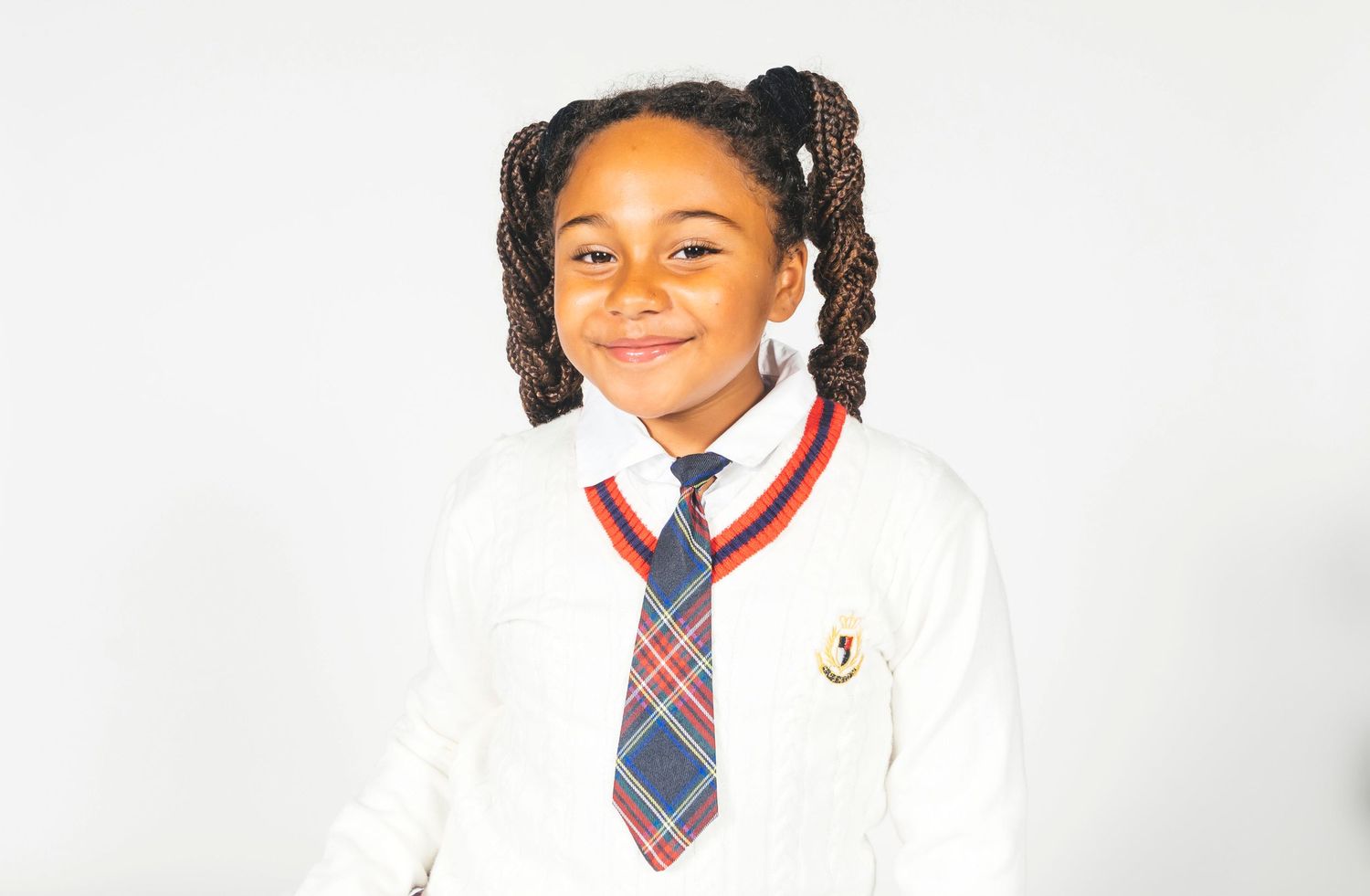 Young girl in school uniform with plaid tie and white blazer, smiling confidently against white background.