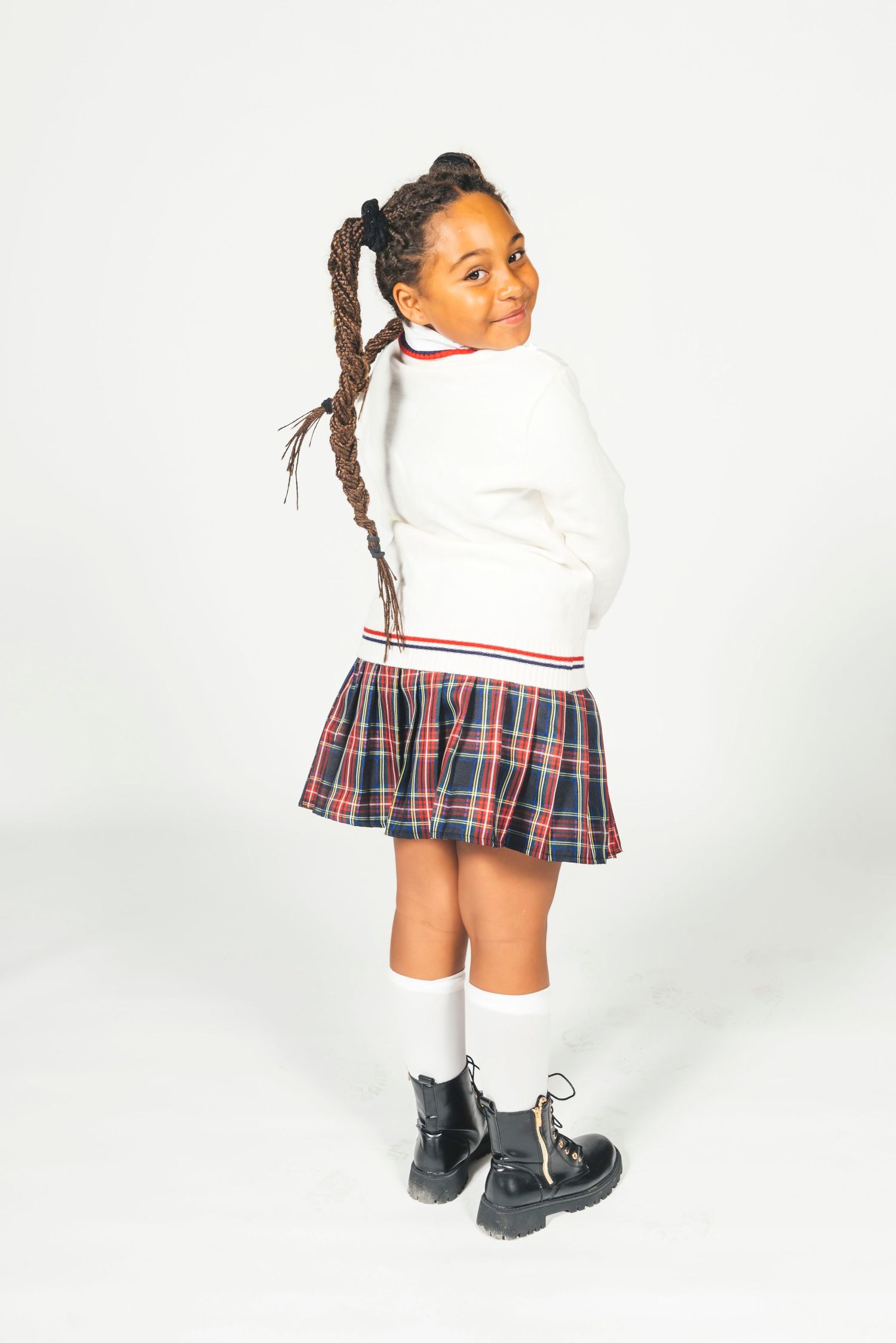 Young girl in school uniform with plaid skirt, white sweater, knee socks, and black boots, smiling over shoulder.