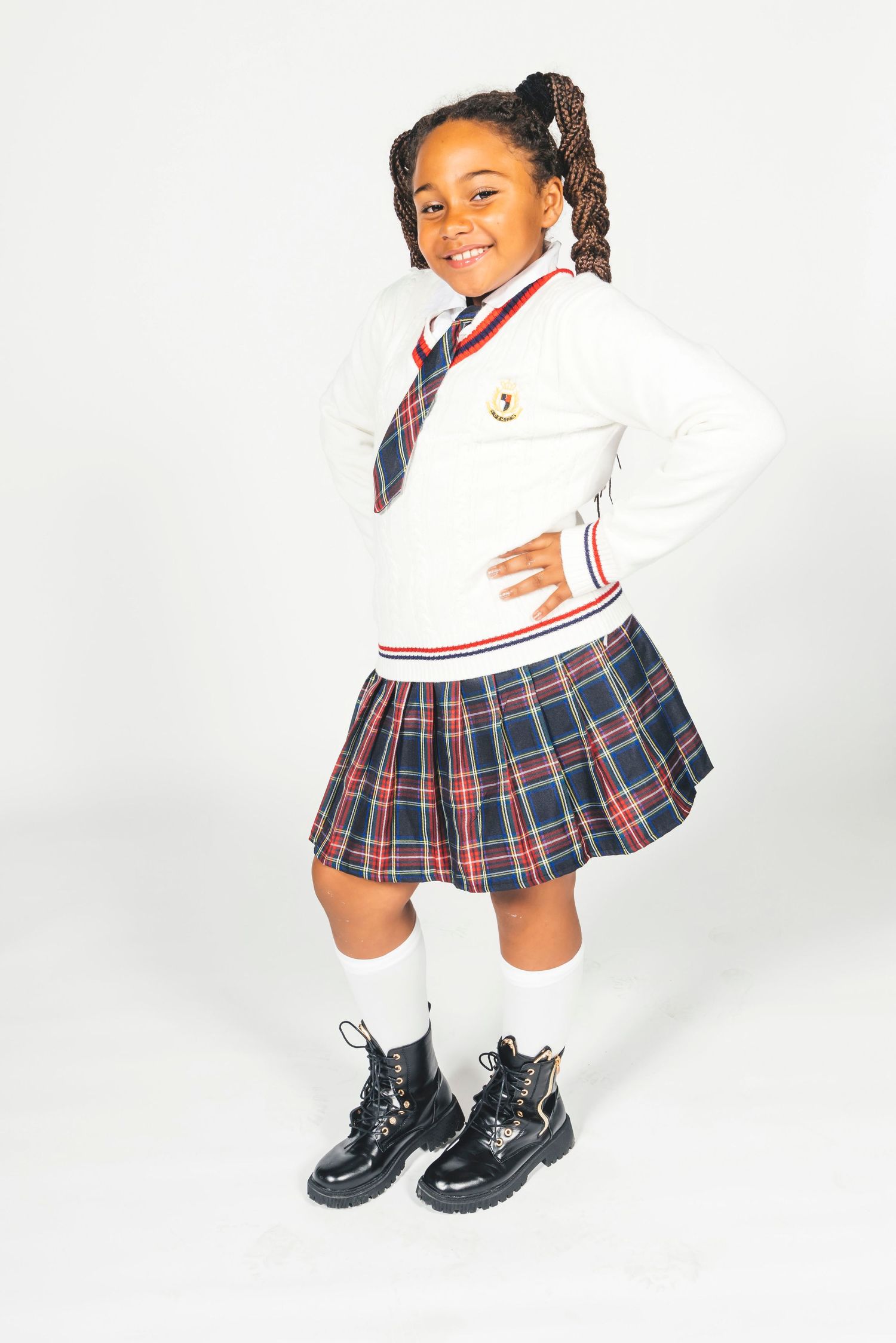 Young girl in school uniform with plaid skirt, white sweater, tie, knee socks, and black boots, smiling confidently.