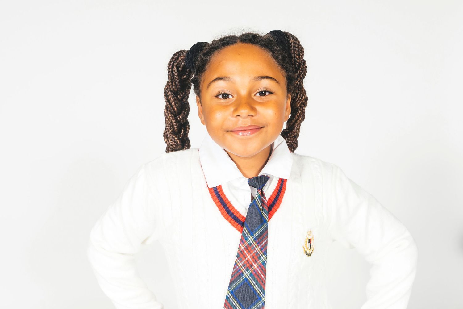 Young girl in school uniform with white cardigan, plaid tie, and braided pigtails smiling confidently.