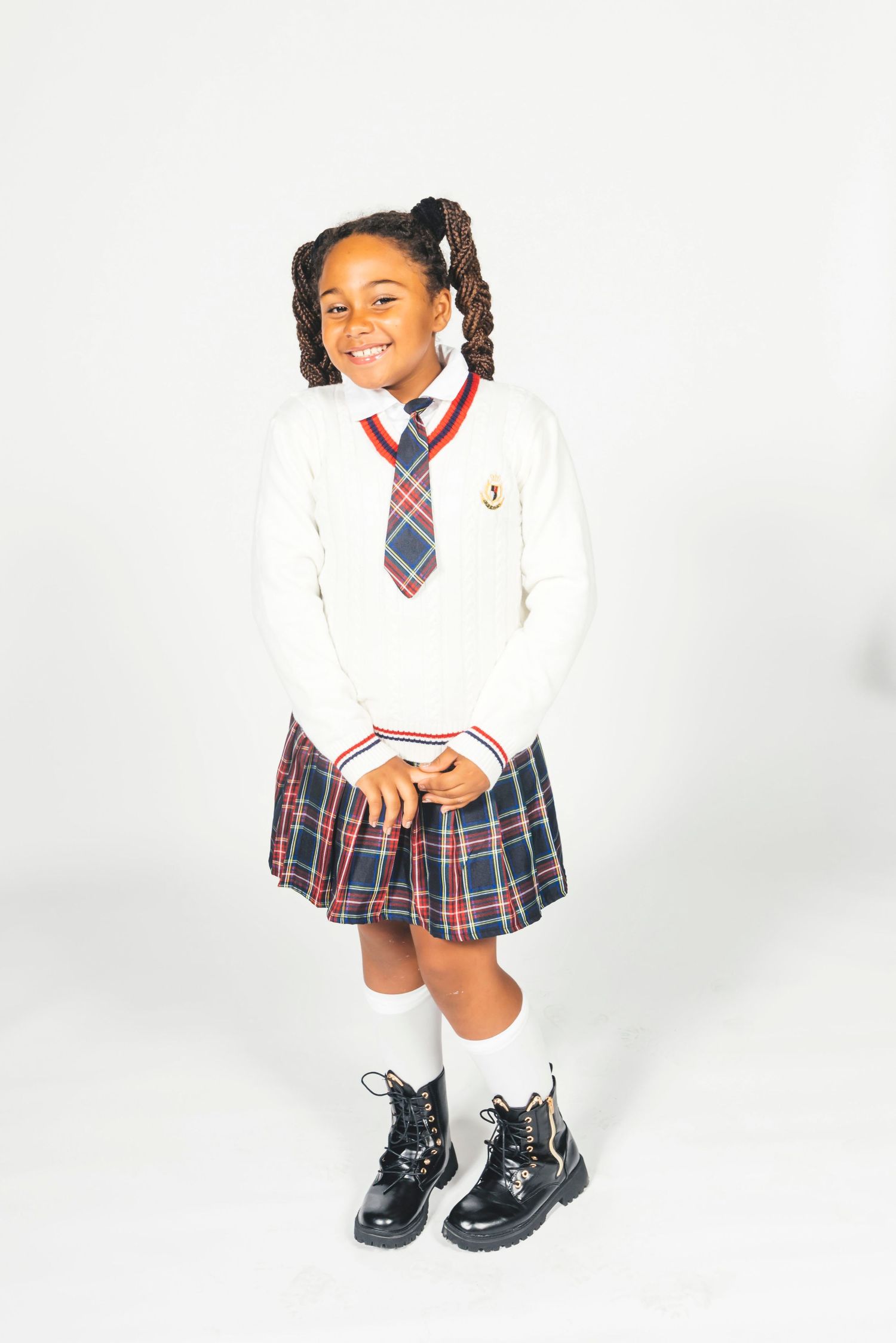 Smiling girl in school uniform with plaid skirt, white sweater, tie, knee socks, and black boots poses on white background.