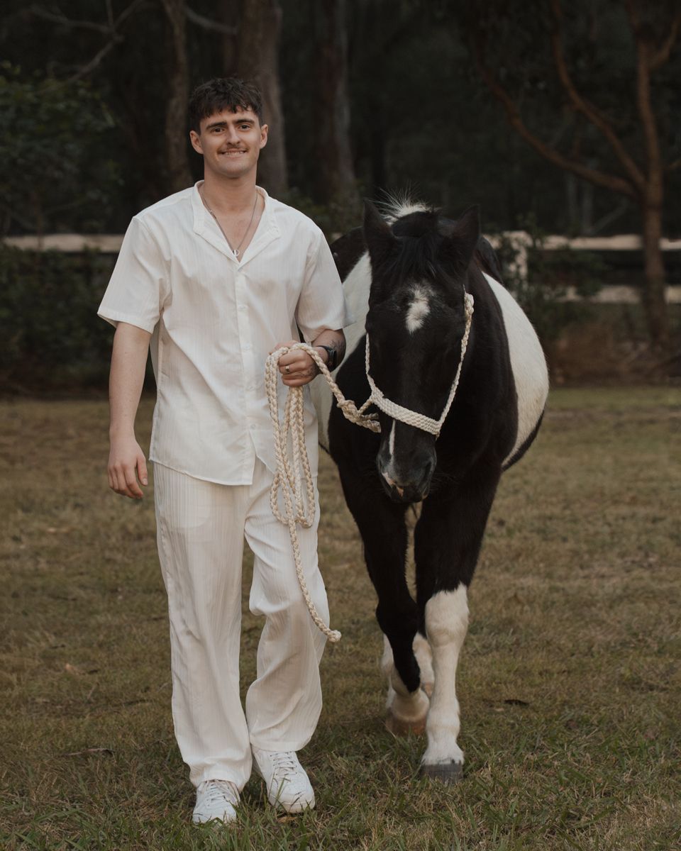 man in white linen outfit holds lead of black and white pony on green grass near trees during a family photoshoot with a horse in Sydney