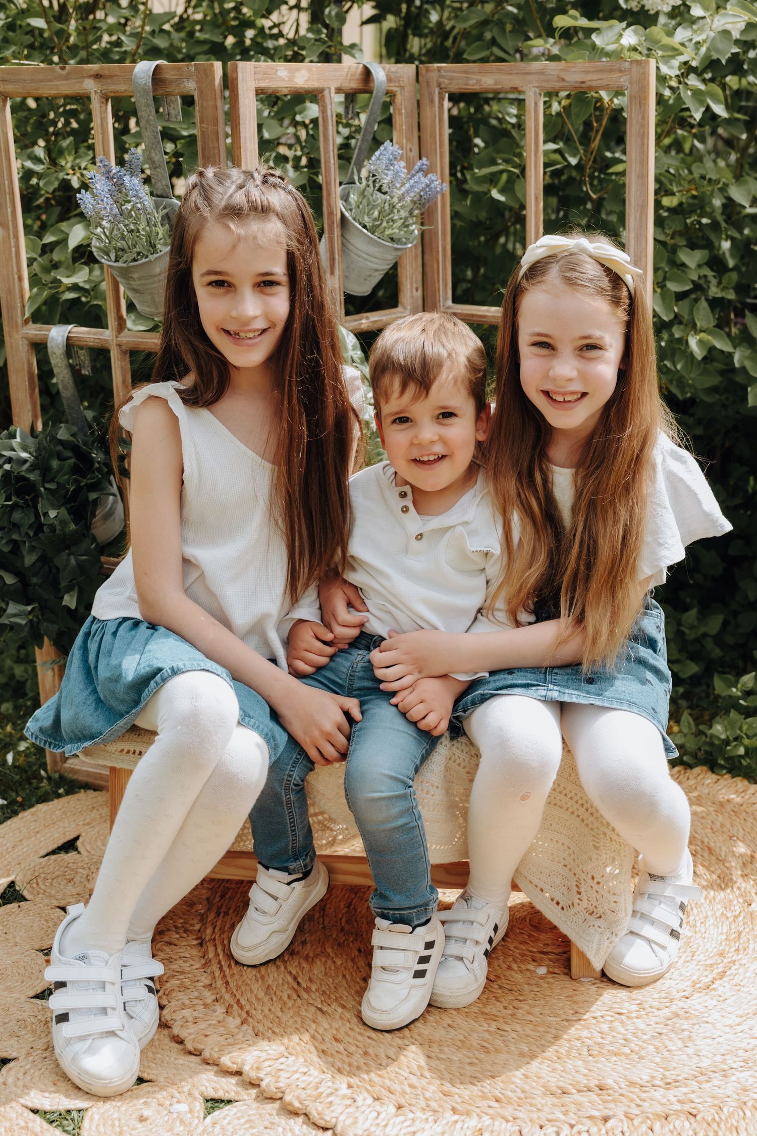 Two girls and a toddler boy sit together on a wooden bench outdoors, wearing white tops and denim outfits.