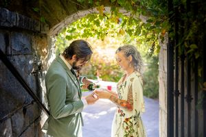 Couple shares champagne under vine-covered stone archway in sunlit garden setting.