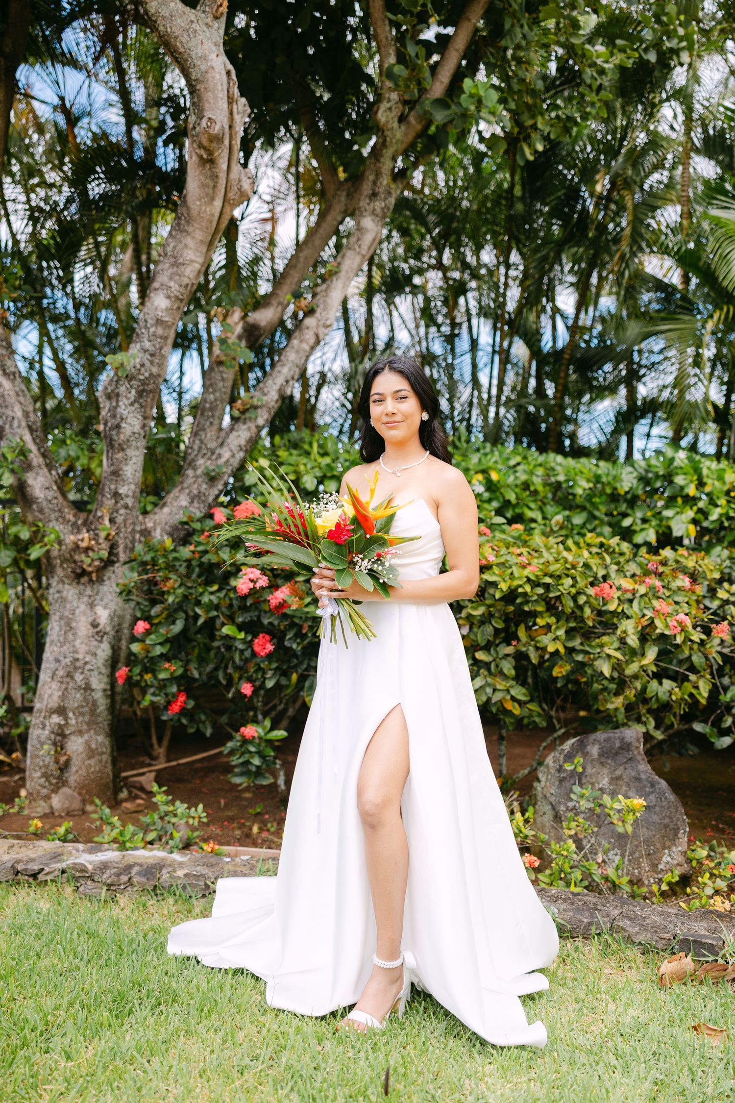 A bride in a white slit gown holds a tropical bouquet in a lush garden setting.