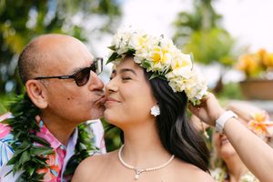 A father kisses his daughter's cheek as she wears a white plumeria flower crown.