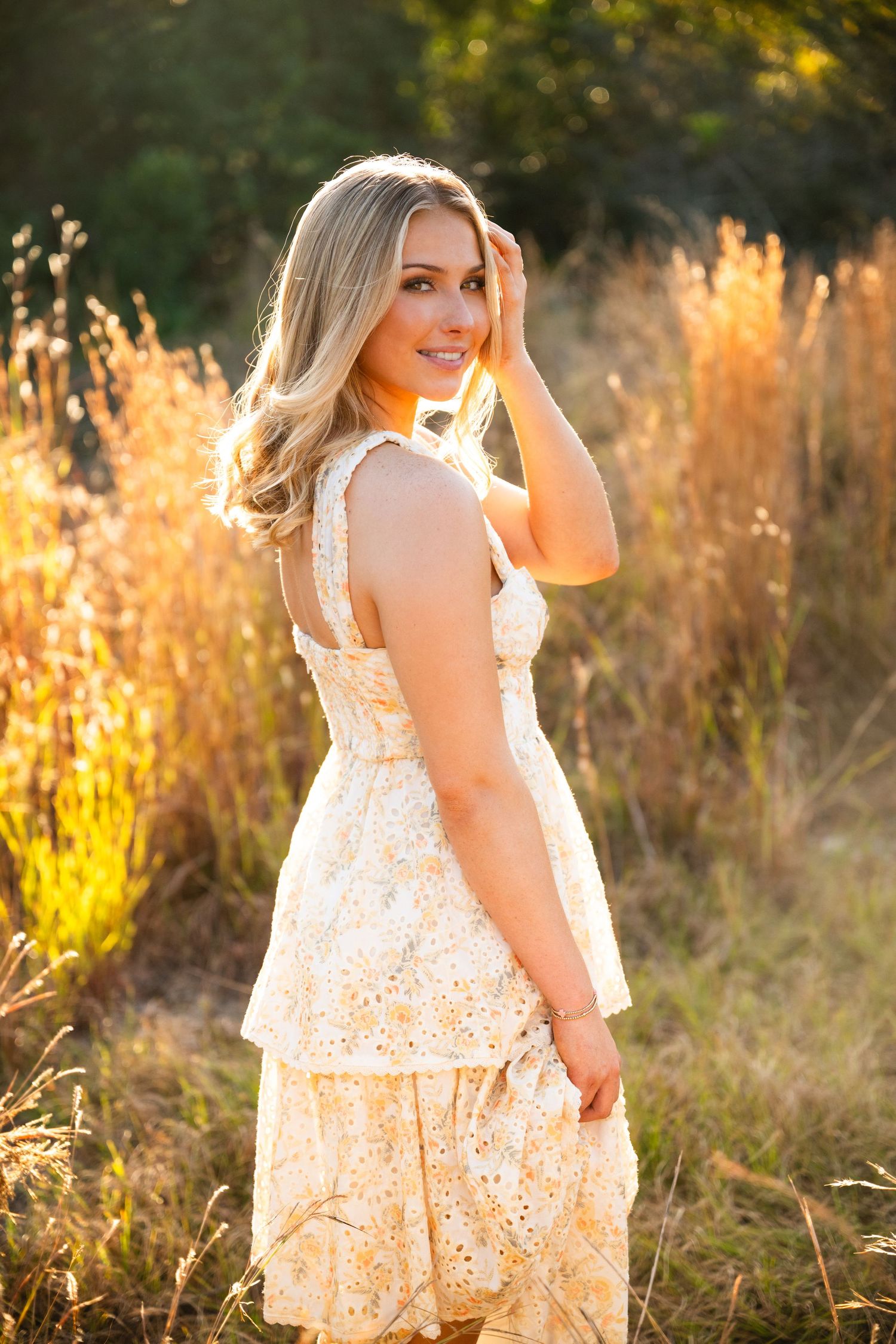 Blonde woman in a floral tiered dress poses in golden sunlit field, smiling over her shoulder.