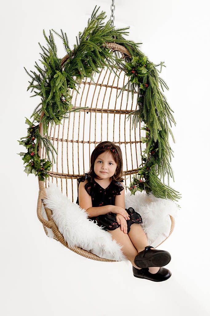 Young girl in black dress sitting in a wicker hanging egg chair decorated with Christmas greenery and holly berries.
