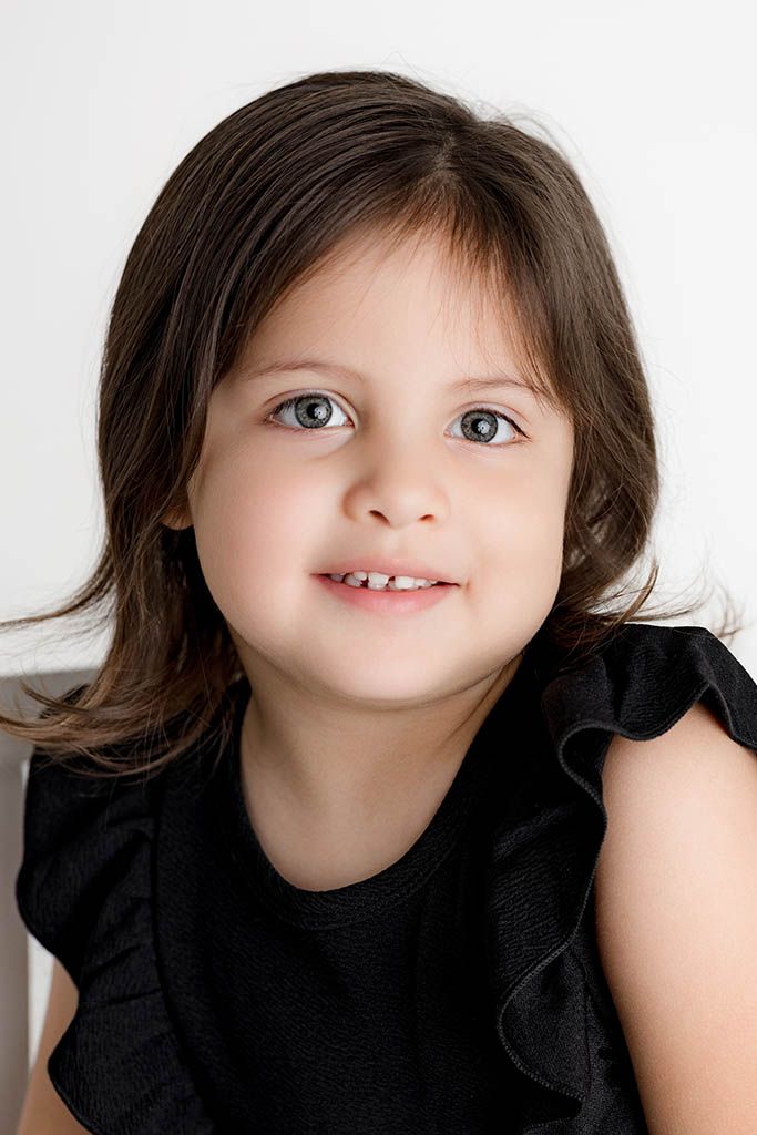 Cute young girl with dark hair and blue eyes smiling, wearing a black ruffled dress against a white background.