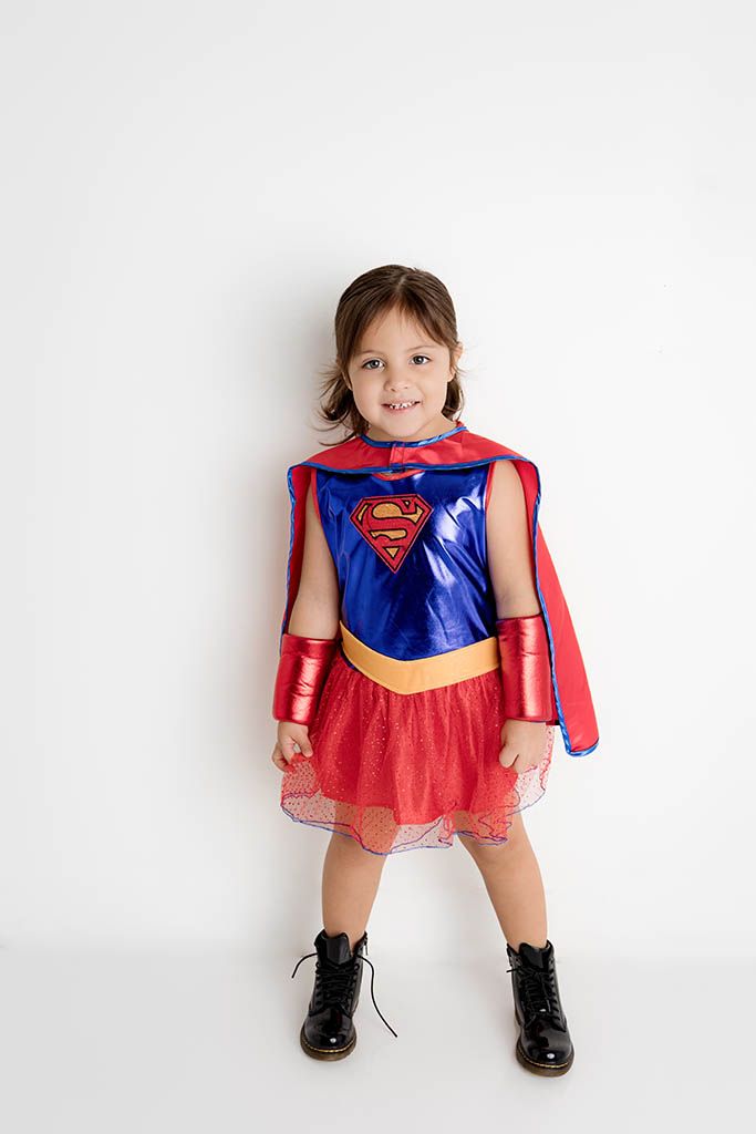Young girl wearing a Supergirl costume with blue top, red cape, tutu skirt, and black boots, smiling against white background.