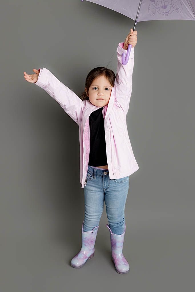 Young girl in pink raincoat, jeans, and purple rain boots holds umbrella overhead with arms outstretched on gray background.
