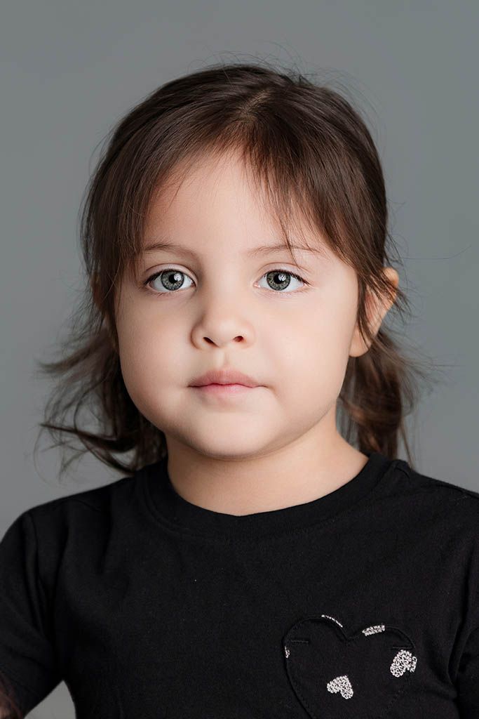 A young girl with dark hair and blue-gray eyes wearing a black t-shirt, posing for a portrait against a gray background.