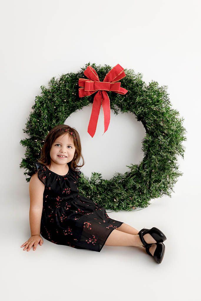 Young girl in black floral dress sitting in front of large Christmas wreath with red bow on white background.