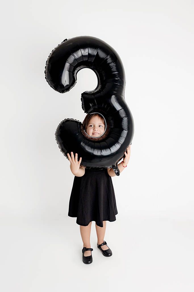 A young girl in a black dress holds a large black number 3 balloon in front of her face on a white background.