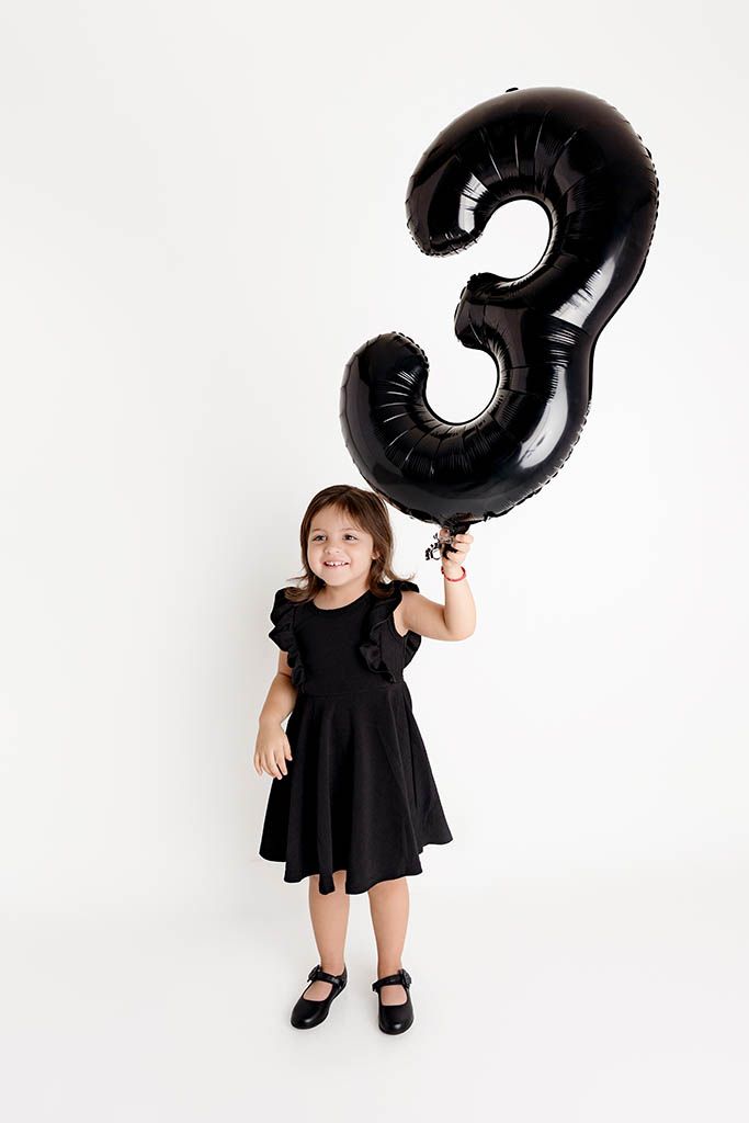 A smiling little girl in a black dress holds a large black number 3 balloon against a white background.
