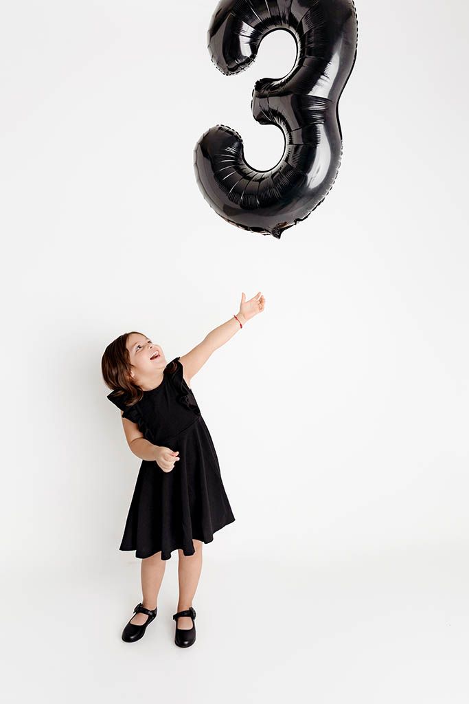 A young girl in a black dress reaches up toward a black number 3 balloon on a white background.