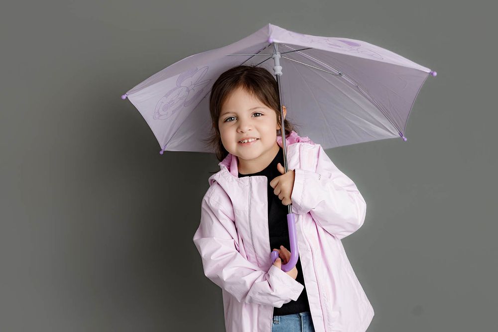 Young girl in pink jacket holding a lavender umbrella against gray background.